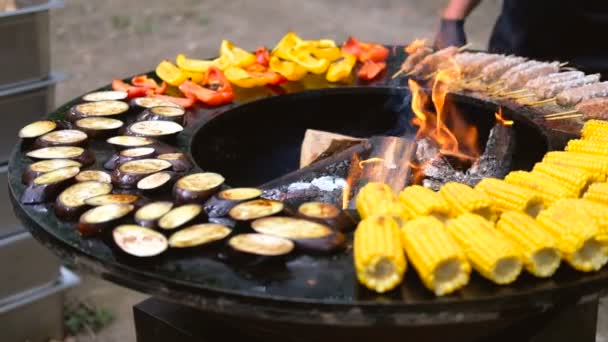 Barbecue rond avec feu ouvert à l'intérieur. Des repas pour le pique-nique d'été sont en préparation : maïs, aubergine, poivron, kebab. Les mains masculines en gants noirs retournent la nourriture avec des pinces pour barbecue . — Video