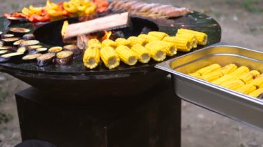 Yuvarlak barbekü ızgara içinde açık ateş ile. Yaz pikniği için yemekler hazırlanıyor: mısır, patlıcan, biber, kebap. Siyah eldivenli erkek eller barbekü maşalarıyla yiyecekleri teslim ediyor.