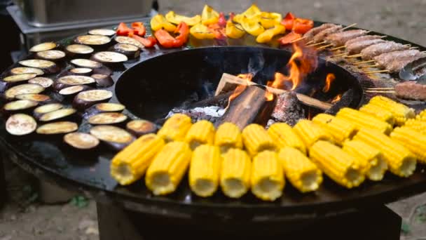 Parrilla redonda con fuego abierto en el interior. Se preparan comidas para el picnic de verano: maíz, berenjena, pimiento, kebab. Las manos masculinas en guantes negros dan vuelta a la comida con pinzas para barbacoa . — Vídeos de Stock