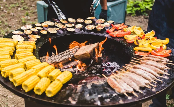 内部に暖炉が付いている円形のバーベキューグリル。夏のピクニックのための食事を準備しています:トウモロコシ、ナス、ピーマン、ケバブ。黒い手袋の男性の手は、バーベキュートングで食べ物をひっくり返します. — ストック写真