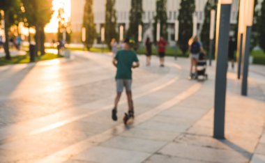 Parktaki ağaçlardan gelen uzun gölgelerle bulanık arka plan. Parkta yürüyen tanınmayan insanların siluetleri.