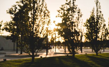 Parktaki ağaçlardan gelen uzun gölgelerle bulanık arka plan. Parkta yürüyen tanınmayan insanların siluetleri.