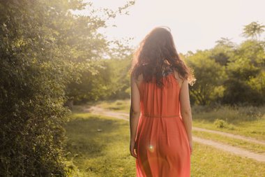 Beautiful young girl with dark curly hair in bright orange dress walks into the distance through forest or field at sunset. Romantic mood. Loneliness.