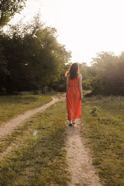 Beautiful young girl with dark curly hair in bright orange dress walks into the distance through forest or field at sunset. Romantic mood. Loneliness. — 图库照片