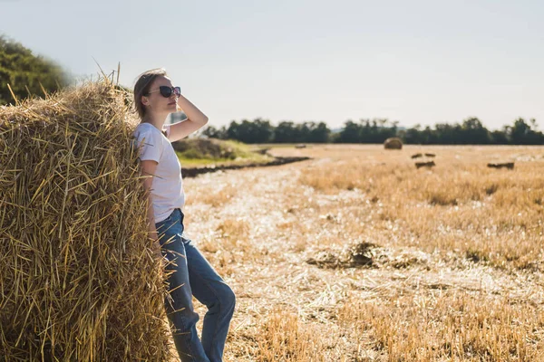 Mladá žena s hnědými vlasy v bílém tričku a modrými džínami pózuje po sklizni na poli u kupky sena. Teplé osvětlení západu slunce. — Stock fotografie