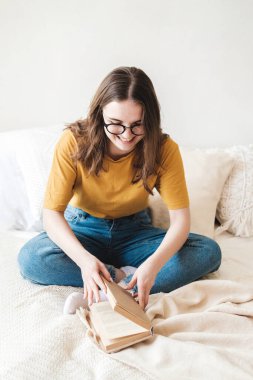 Gözlüklü, turuncu tişörtlü ve kot pantolonlu genç güzel bir kız yatakta yastıklarla oturur ve kağıt kitap okur. Evde eğitim kavramı, hobi..
