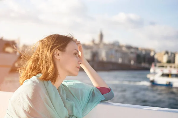 Ung vacker flicka i rosa och blå klänning sitter på båt eller färja vid solnedgången och eftertänksamt tittar på havet. — Stockfoto
