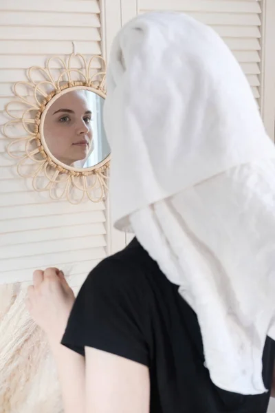 Junge schöne Frau mit einem Handtuch auf dem Kopf nach der Dusche im Badezimmer betrachtet ihr Spiegelbild im Spiegel an der Wand. Konzept weiblicher Haut- und Haarpflege am Morgen. — Stockfoto