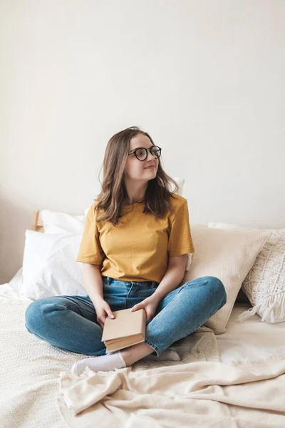 Die junge hübsche Studentin mit Brille, orangefarbenem T-Shirt und blauer Jeans sitzt auf einem Bett mit Kissen und liest ein Buch. Selbstbildungskonzept zu Hause während der Quarantäne. Hausaufgabenbetreuung, Hobby. — Stockfoto