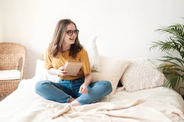 Gözlüklü, turuncu tişörtlü ve kot pantolonlu genç güzel bir kız yatakta yastıklarla oturur ve kağıt kitap okur. Karantina sırasında evde kendi kendine eğitim kavramı. Evde eğitim, hobi..