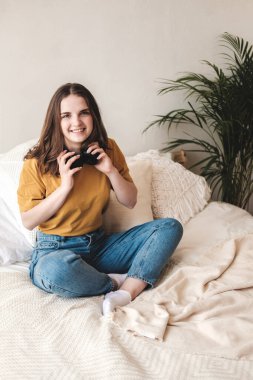 Turuncu tişörtlü ve mavi kotlu genç bir kız öğrenci yastıklarla yatağa oturur ve bir sesli kitap, müzik ve kulaklıkla podcast dinler. Karantina sırasında evde eğitim kavramı.