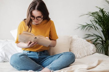 Gözlüklü, turuncu tişörtlü ve kot pantolonlu genç güzel bir kız yatakta yastıklarla oturur ve kağıt kitap okur. Karantina sırasında evde kendi kendine eğitim kavramı. Evde eğitim, hobi..