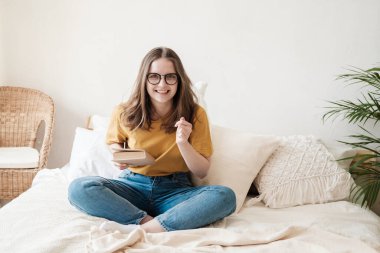 Young beautiful girl student in glasses, an orange T-shirt and blue jeans sits on bed with pillows and reads paper book. Self-education concept at home during quarantine. Home schooling, hobby. clipart