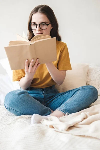 Gözlüklü, turuncu tişörtlü ve kot pantolonlu genç güzel bir kız yatakta yastıklarla oturur ve kağıt kitap okur. Karantina sırasında evde kendi kendine eğitim kavramı. Evde eğitim, hobi.. — Stok fotoğraf