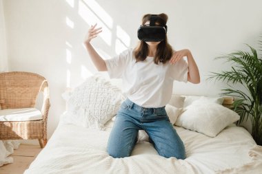 Beyaz tişörtlü ve kot pantolonlu genç güzel bir kadın oyun oynuyor, yatakta sanal gerçeklik kulaklığıyla dans ediyor. Eğlence anlayışı, karantina sırasında evde öğrenme.