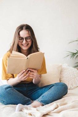 Gözlüklü, turuncu tişörtlü ve kot pantolonlu genç güzel bir kız yatakta yastıklarla oturur ve kağıt kitap okur. Karantina sırasında evde kendi kendine eğitim kavramı. Evde eğitim, hobi..