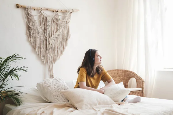 Turuncu tişörtlü ve mavi kotlu genç bir kız öğrenci yastıklarla yatağa oturur ve bir sesli kitap, müzik ve kulaklıkla podcast dinler. Karantina sırasında evde eğitim kavramı.