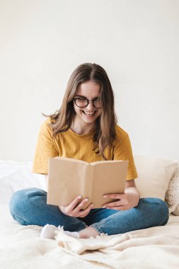 Gözlüklü, turuncu tişörtlü ve kot pantolonlu genç güzel bir kız yatakta yastıklarla oturur ve kağıt kitap okur. Karantina sırasında evde kendi kendine eğitim kavramı. Evde eğitim, hobi..