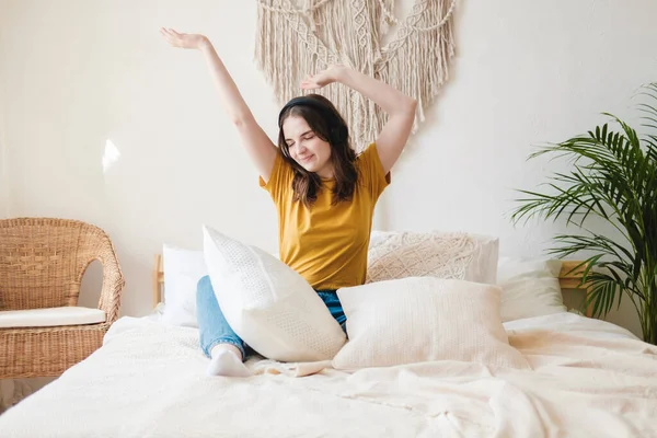 Turuncu tişörtlü ve mavi kotlu genç bir kız öğrenci yastıklarla yatağa oturur ve bir sesli kitap, müzik ve kulaklıkla podcast dinler. Karantina sırasında evde eğitim kavramı.