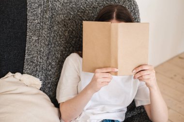 Kot pantolonlu ve beyaz tişörtlü genç bir kadın gri koltukta oturur ve kağıt kitap okur. Rahatlatıcı tatil kavramı, ev konforu, kızlık zarı, yavaş yaşam.