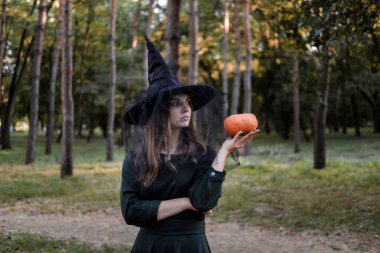Siyah elbiseli ve cadı şapkalı genç bir kadın elinde bir balkabağı tutuyor. Cadılar Bayramı partisi kostümü. Orman, sonbahar ağaçlarının olduğu park.
