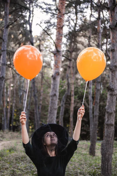 身穿深色衣服头戴黑色帽子的年轻漂亮女子手里拿着橙色气球。万圣节派对服装。森林，有秋天树的公园. — 图库照片