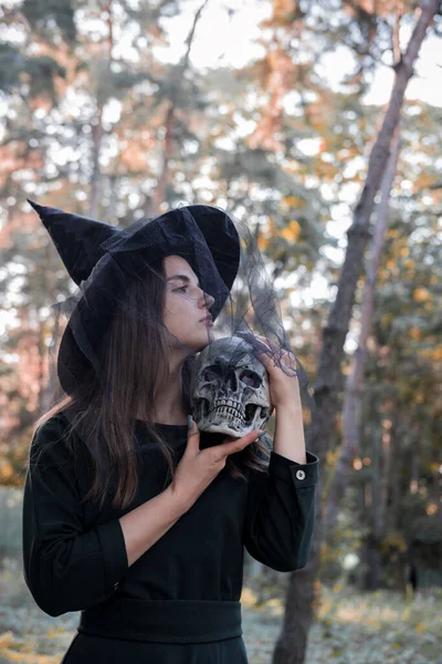 Junge hübsche Frau in dunklem Kleid und Hexenhut hält einen Totenkopf in ihren Händen. Halloween-Party-Kostüm. Wald, Park mit Herbstbäumen. — Stockfoto