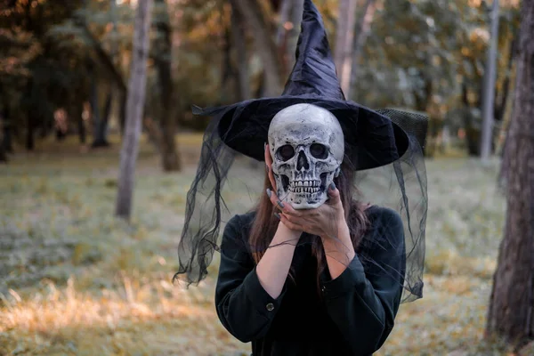 Jeune belle femme mignonne en robe sombre et chapeau de sorcières tient un crâne dans ses mains. Costume de fête d'Halloween. Forêt, parc avec arbres d'automne. — Photo