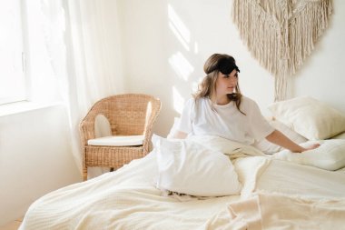 Genç ve güzel bir kadın rahat bir yatakta uzanıyor ve sabahları rahat bir evde uyumak için maske takıyor. Kendini izole etmek, karantina ve salgın sırasında evde kalmak..