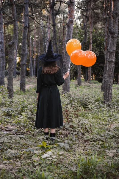 Jeune jolie femme mignonne en robe sombre et chapeau de sorcières tient des ballons orange dans ses mains. Costume de fête d'Halloween. Forêt, parc avec arbres d'automne. — Photo