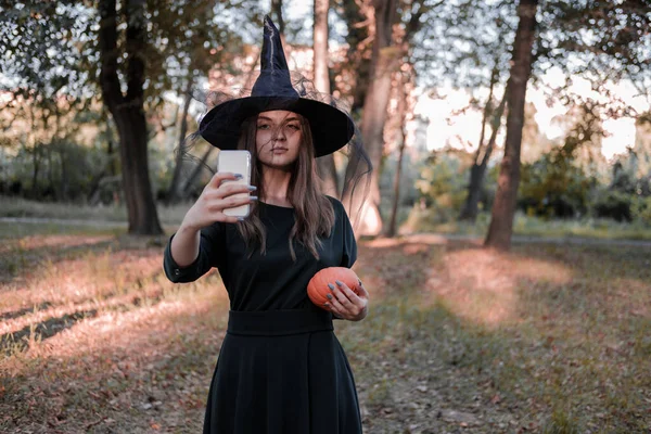 Jeune jolie femme mignonne en robe sombre et chapeau de sorcières fait un selfie avec citrouille pour les réseaux sociaux. Costume de fête d'Halloween. Forêt, parc avec arbres d'automne. — Photo