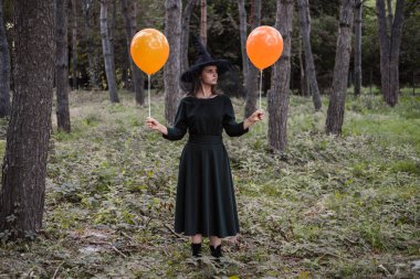 Young cute beautiful woman in dark dress and witchs hat holds orange balloons in her hands. Halloween party costume. Forest, park with autumn trees. clipart