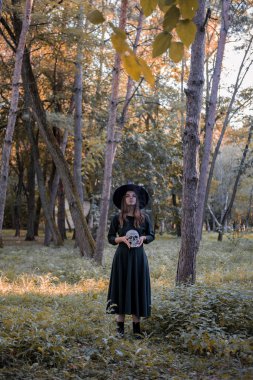 Siyah elbiseli ve cadı şapkalı güzel bir kadın elinde bir kafatası tutuyor. Cadılar Bayramı partisi kostümü. Orman, sonbahar ağaçlarının olduğu park.