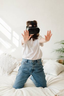 Beyaz tişörtlü ve kot pantolonlu genç güzel bir kadın oyun oynuyor, yatakta sanal gerçeklik kulaklığıyla dans ediyor. Eğlence anlayışı, karantina sırasında evde öğrenme.