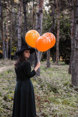 Koyu renk elbiseli ve şapkalı güzel bir kadın ellerinde turuncu balonlar tutuyor. Cadılar Bayramı partisi kostümü. Orman, sonbahar ağaçlarının olduğu park.