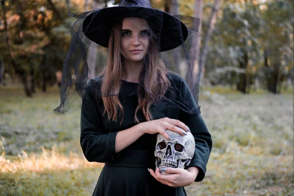 Jeune belle femme mignonne en robe sombre et chapeau de sorcières tient un crâne dans ses mains. Costume de fête d'Halloween. Forêt, parc avec arbres d'automne. — Photo