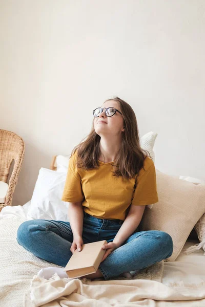 Ung vacker flicka student i glasögon, en orange T-shirt och blå jeans sitter på sängen med kuddar och läser pappersbok. Självskolekoncept hemma under karantänen. Hemundervisning, hobby. — Stockfoto