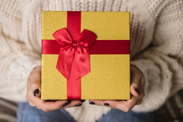 Mujer joven se sienta en la cama en un acogedor suéter blanco de lana de punto y la celebración de la caja de regalo con oro rojo. Hygge, Año Nuevo, Navidad, preparativos navideños. Velas, bolas de Navidad, regalos, malvaviscos de cacao. — Foto de Stock