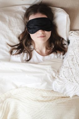 Genç ve güzel bir kız rahat bir yatakta uzanıyor. Sabahları rahat bir odada uyku maskesi takıyor. Evde dinlenme, uyku, uykusuzluk, kendini izole etme, karantina sırasında evde kalma, salgın hastalık.