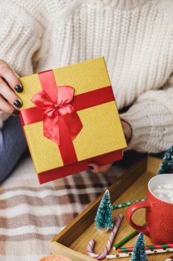 Genç bir kadın rahat örülmüş yün beyaz bir kazakla yatağında oturuyor ve elinde kırmızı altınla hediye kutusu tutuyor. Hygge, yeni yıl, Noel. Marşmelovlu bir kupa kakao, oyuncak ağaç, mum ve yıldızlı tahta tepsi..