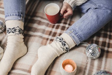 Genç bir kadın rahat örülmüş yün beyaz bir kazakla ekose üzerinde oturuyor ve çoraplar ellerinde bir fincan kakao tutuyor. Hygge Yeni Yıl, rahat bir Noel, tatil hazırlıkları. Mumlar, Noel baloları.