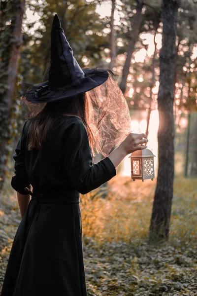 Mladá žena v tmavých šatech a čarodějnickém klobouku drží lucernu se svíčkami v rukou a osvětluje les. Halloween mejdan kostým. Park s podzimními stromy. — Stock fotografie