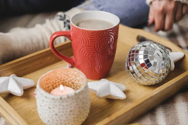 Young woman sits on plaid in cozy knitted woolen white sweater and holds cup of cocoa in her hands. Hygge wooden tray with mug of chocolate, toy tree, candle, stars. — Stock Photo, Image