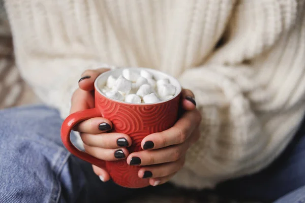 Young woman sits on plaid in cozy knitted woolen white sweater and holds cup of cocoa with marshmallows in her hands. Hygge New Year, cozy Christmas, preparation for holidays. — Stock Photo, Image