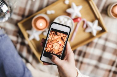 Akıllı telefonlu bir kadın sosyal ağlar için yılbaşı resmi ya da Noel resmi çekiyor. Tatil hazırlığı, içerik yaratmak. Kakao kupalı ahşap tepsi lokumlar, mum, yıldızlar.