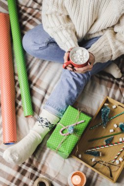 Genç bir kadın, rahat, yün, beyaz bir kazakla ekose üzerinde oturur ve elinde lokum dolu bir fincan kakao tutar. Hygge Yeni Yıl, tatil hazırlıkları. Mumlar, Noel baloları, hediyeler, ambalaj kağıtları..