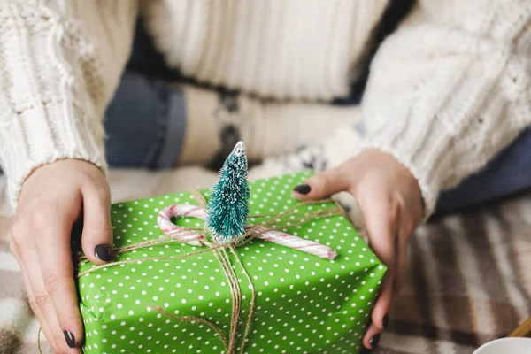 Молода жінка сидить на плетеному в'язаному вовняному білому светрі, шкарпетках і обгортає різдвяний подарунок у папері для обгортання крапок. Дерев'яний піднос з чашкою какао з зефіром, іграшковим деревом, свічкою, соломою . — стокове фото