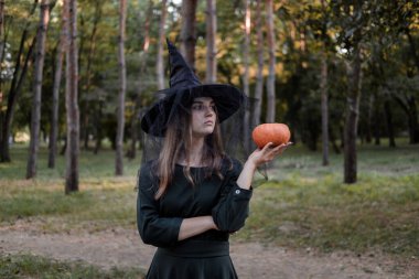 Siyah elbiseli ve cadı şapkalı genç bir kadın elinde bir balkabağı tutuyor. Cadılar Bayramı partisi kostümü. Orman, sonbahar ağaçlarının olduğu park.
