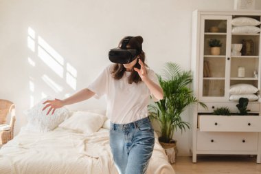 Kot pantolonlu ve beyaz tişörtlü genç bir kadın sanal gerçeklik kaskı giyiyor, oyun oynuyor, çalışıyor, yan odada video izliyor. Modern teknoloji kavramı, VR, artırılmış sanal oyunlar, eğlence.