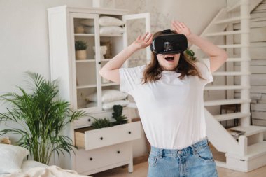 Kot pantolonlu ve beyaz tişörtlü genç bir kadın sanal gerçeklik kaskı giyiyor, oyun oynuyor, çalışıyor, yan odada video izliyor. Modern teknoloji kavramı, VR, artırılmış sanal oyunlar, eğlence.